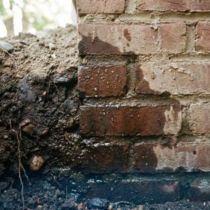 Doorsnede bakstenen funderingsmuur met vocht dat omhoog sijpelt uit verzadigde grond, waterdruppels op metselwerk zichtbaar