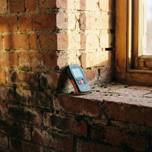 Vochtmeter tegen bakstenen muur met waterschade, overgang van donkere vlekken onderaan naar droge stenen bovenaan