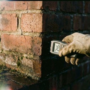 Vochtmeter meet waterschade op bakstenen muur met donkere vlekken en scheuren in voegen