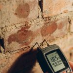 Professionele vochtmeter tegen bakstenen muur toont verschillende vochtniveaus, digitaal display zichtbaar op handheld apparaat