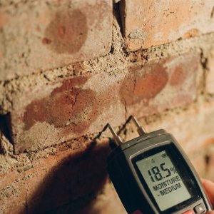 Professionele vochtmeter tegen bakstenen muur toont verschillende vochtniveaus, digitaal display zichtbaar op handheld apparaat