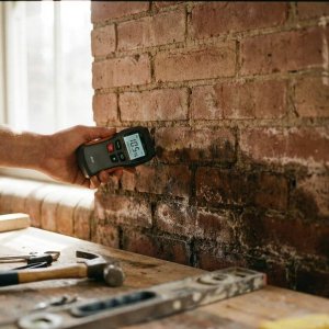 Vochtmeter toont normale waarden tegen bakstenen muur met waterschade onderaan en droge metselwerk bovenaan