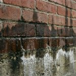 Bakstenen muur met vochtschade en watervlekken die omhoog klimmen vanaf betonnen fundering, condensdruppels zichtbaar