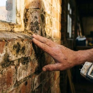 Bouwvakker onderzoekt waterlek en schimmel op bakstenen muur met vochtmeter, condensdruppels zichtbaar in gouden licht