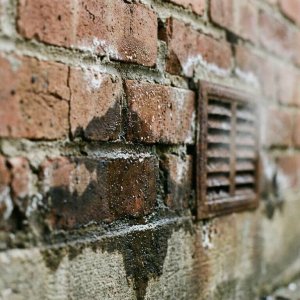 Bakstenen muur met waterschade en vochtplekken die omhoog kruipen vanaf de fundering, met ventilatierooster