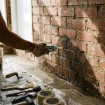 Bouwvakker past witte waterdichte kit toe op vochtige bakstenen muur met vochtplekken, gereedschap op zeil eronder