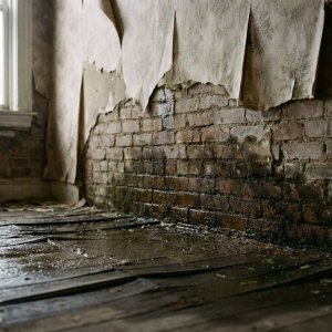 Waterschade aan bakstenen muur met donkere vochtplekken, vervormde houten vloer en loslaten behang door vocht