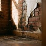 Waterschade aan bakstenen muur met afbladderende verf, vocht en vervormde houten vloerplanken in hoek