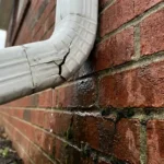 Beschadigde witte kunststof regenpijp met scheur lekt water op bakstenen fundering, donkere vochtschade zichtbaar op muur