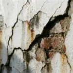 Gebarsten witte stucmuur met vochtplekken en zichtbare bakstenen door grote scheuren