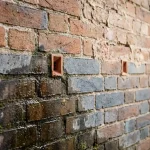 Verweerde bakstenen muur met vochtplekken en droge gedeelten, keramische ventilatie-elementen in voegen, natuurlijk daglicht