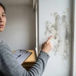 Bezorgde huurder wijst naar donkere vochtplekken op witte appartementmuur, met inspectieformulieren op houten tafel