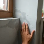 Hand raakt vochtige muur aan met watervlekken en verkleuring in Nederlandse woonkamer hoek