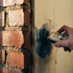 Doorsnede van bakstenen muur met waterdruppels buiten en vochtmeter die binnenvochtvlekken meet