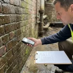 Professionele bouwinspecteur onderzoekt vochtige bakstenen muur met digitale vochtmeter, druppels zichtbaar op rode baksteen