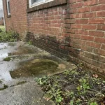 Rode bakstenen huismuur met donkere vochtplekken bij de fundering en wateroverlast op betonnen terras