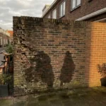 Verweerde bakstenen muur met vochtschade en donkere watervlekken die omhoog kruipen vanaf de fundering