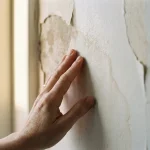Homeowner raakt vochtschade op witte muur aan met donkere watervlekken en loslaten behang