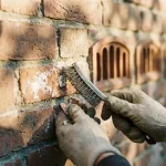 Handen met werkhandschoenen verwijderen witte zoutuitslag van verweerde bakstenen muur met staalborstel