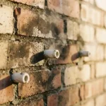 Vochtige bakstenen muur met witte keramische ventilatiepijpjes en opstijgende waterdamp in warm middaglicht