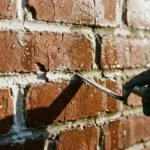 Verweerde bakstenen muur met afbrokkelende witte voegmortel, waterdruppels en professioneel gereedschap dat beschadigde voegen onderzoekt