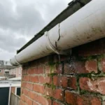 Beschadigde witte dakgoot met scheur lekt water op natte rode bakstenen muur met donkere watervlekken en mos