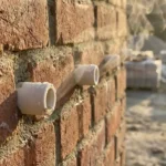 Doorsnede van bakstenen muur met witte keramische buizen, waterdamp stijgt op uit openingen in warm zonlicht