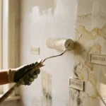 Verfroller brengt witte primer aan op waterbeschadigde muur met zichtbare vlekken en CAREBRICK ventilatie-elementen