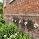 Verweerde bakstenen huis met donkere vochtplekken en witte keramische ventilatiebuizen langs de onderste muur