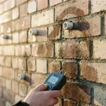 Vochtmeter meet vochtniveaus op bakstenen muur met ventilatieopeningen en vochtplekken