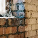 Bakstenen muur in doorsnede met vochtdruppels en donkere vlekken die overgaan naar droge gebieden, zonlicht creëert verdampingsnevel