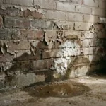 Verweerde bakstenen muur met donkere watervlekken, witte zoutafzettingen en afbladderende verf boven betonnen vloer