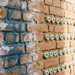Bakstenen muur met vochtplekken links en witte keramische buizen in voegen rechts, natuurlijk daglicht toont contrast