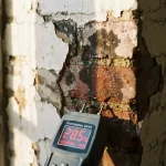 Doorsnede van bakstenen muur met donkere watervlekken en vochtschade onder afbladderende witte verf, vochtmeter toont zorgwekkende metingen