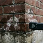 Bakstenen muurhoek met vochtschade, donkere watervlekken, witte zoutafzettingen en professionele vochtmeter
