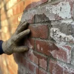 Hand in werkhandschoen onderzoekt vochtschade op bakstenen muur met donkere watervlekken en witte zoutafzettingen