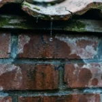 Waterdruppel valt van verweerde dakpan op vochtige bakstenen muur met zichtbare waterschade
